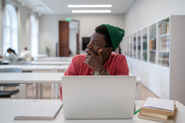 Procrastination concept. Thoughtful Black student man looking away while studying in library,...