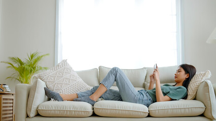 Happy young asian woman relaxing at home. Female is lying down on sofa and using mobile smartphone