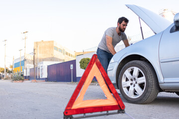 young man inspecting the engine of his car, hazard triangle on the road, car insurance concept