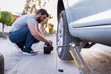 young man loosening a nut of his car tire, changing a flat tire on the road, car jack being used