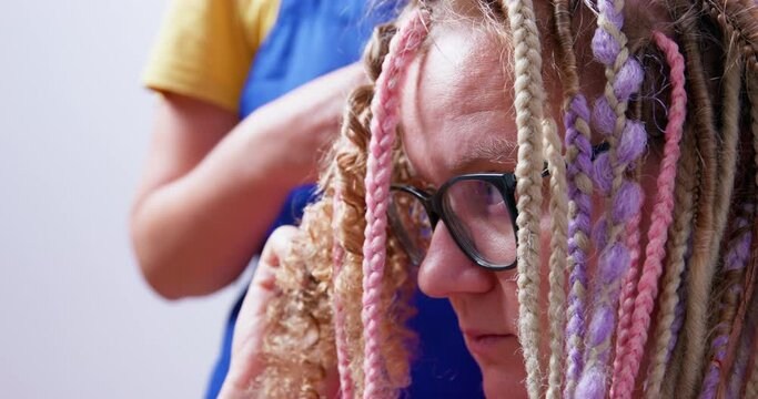 Young blonde woman with glasses sits and holds her curls while master unwinds her multicolored box braids, side view, close up. Different female hairstyles for long hair. Time to change style.