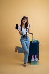 Full length of young attractive Asian woman traveler in casual clothes wearing sunglasses with luggage, camera, snorkel and flippers showing smart mobile phone for presenting