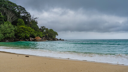Rainy foggy day on a tropical island. The waves of the turquoise ocean are foaming on the sand of the beach. A hill overgrown with green vegetation, against a cloudy sky. Boulders in the water.
