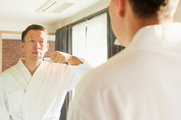 Asian man in white bathrobe brushing teeth standing in front of mirror, oral care concept.