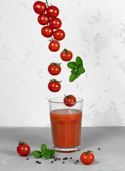 Small red cherry tomatoes fly into a glass with tomato juice on a light background.