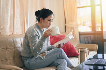 Fototapeta na wymiar woman relaxing at home , woman reading book and drink coffee holding a cup of coffee sitting on sofa - relaxing in her living room reading book on holiday is favorite hobby in home and comfort