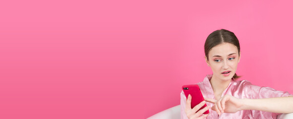 a happy girl with a magenta smartphone in her hands lies in a bathtub filled with inflatable pink balloons on pink background Space for text on the left