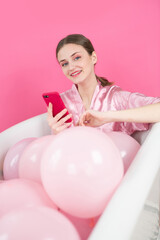 a happy girl with a magenta smartphone in her hands lies in a bathtub filled with inflatable pink balloons on pink background Close up