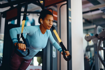 Black sportswoman doing TRX exercises with suspension straps in gym.