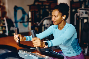 Fototapeta na wymiar Smiling black sportswoman working out on exercise bicycle in gym.