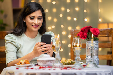 happy smiling girl texting to boyfriend on mobile phone for candle light dinner at home - concept of online dating, valentines day planning and