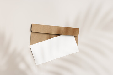 Blank paper card mock up and craft envelope on beige background and palm leaf shadow. White empty invitation on table. Minimal fashion lifestyle photo. Summer Flat lay, top view, copy space.