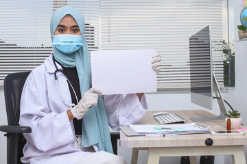 Fototapeta na wymiar Muslim doctor in hijab wearing protective mask and gloves, showing a blank sheet of paper at clinic.