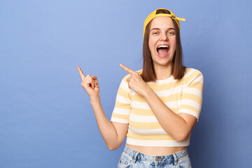 Obraz na płótnie Canvas Photo of extremely happy amazed teenager girl wearing striped T-shirt and baseball cap, pointing at empty wall, showing copy space, advertisement area, posing isolated over blue background,