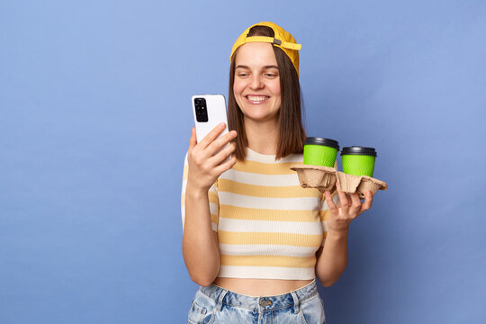 Indoor Shot Of Optimistic Charming Teenager Girl Wearing Striped T-shirt And Baseball Cap Posing Isolated Over Blue Background, Holding Double Coffee To Go And Using Mobile Phone.