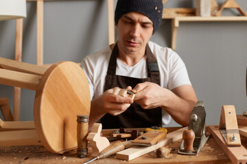 Indoor shot of handsome carpenter making a handmade wooden toy in a home workshop, enjoying wood...