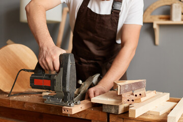Portrait of faceless anonymous carpenter working on woodworking machines in carpentry shop, holding jigsaw in hands, standing at workbench, using instrument.