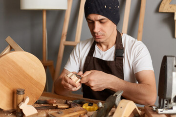 Portrait of carpenter man in brown apron making a handmade wooden toy in a home workshop, handyman...