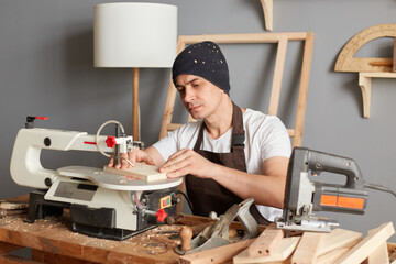 Indoor shot of attractive man carpenter wearing brown apron and black cap, works with wood, using electric jigsaw for making patterns on a wooden block, working in his joinery.