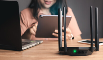 Selective focus at router. Internet router on working table with blurred happy woman using tablet at the background. Fast and high speed internet connection from fiber line with LAN cable connection. - obrazy, fototapety, plakaty
