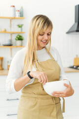 A pastry chef girl in a beige apron is standing in a white kitchen, whipping cream for sweets with a hand whisk in a white plate