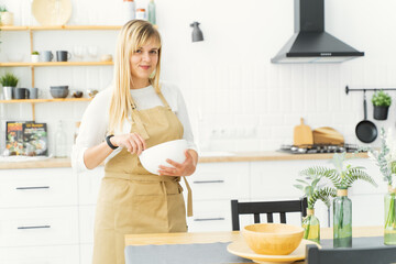 A pastry chef girl in a beige apron is standing in a white kitchen, whipping cream for sweets with a hand whisk in a white plate