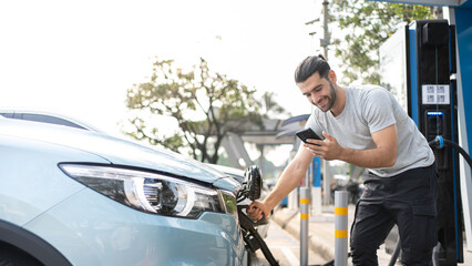 Fototapeta na wymiar Smiling Caucasian man in casual wear Using phone while Charging on his electric car, standing on the charging station and using application. Electric car charging concept.