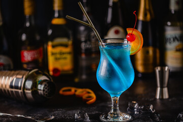Glass of blue lagoon cocktail decorated with orange and cherry at festive bar counter background.