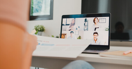 Young Asia businesswoman using laptop talk to colleagues about plan in video call meeting while work from home at living room. Self-isolation, social distancing, quarantine for coronavirus prevention.
