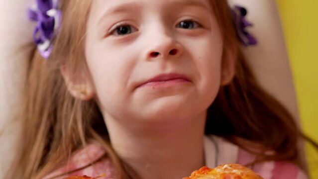 A happy little girl takes a bite of pizza, chews, looks into the frame.