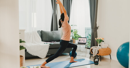 Young attractive Asian woman in sportswear site on mat in the floor virtual fitness class with group of people on a video conference on laptop practice yoga for beginner in living room at home.