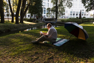 Fototapeta na wymiar An elderly man in a white shirt is sitting on a blanket, on the ground in a park and reading an interesting book. A pensioner alone is resting in nature, passionate about his hobby