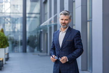 Portrait of successful mature man outside office building, businessman in business suit looking at camera thinking smiling while standing.