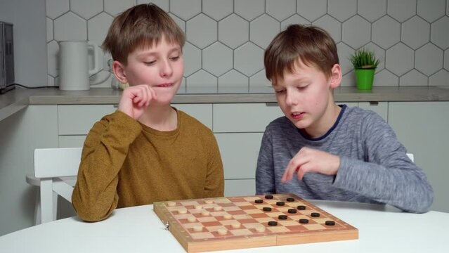 Video of two little smiling, interested, concentrated boys playing checkmates on board on table in kitchen. Gambling and thinking, winner and looser. Strategy board game for brain development