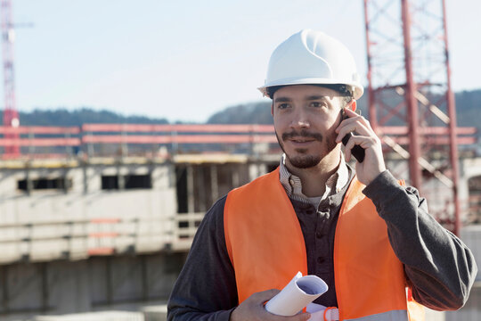 Young male engineer holding blueprint and talking on a mobile phone at construction site, Freiburg Im Breisgau, Baden-Württemberg, Germany - Powered by Adobe