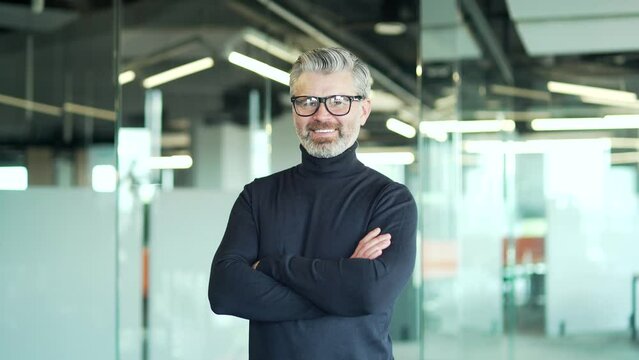 Portrait Of A Happy Mature Gray Haired Bearded Man In Glasses Looking At The Camera While Standing In A Modern Office. Middle Aged Confident Smiling Businessman With A Friendly Look And Crossed Arms