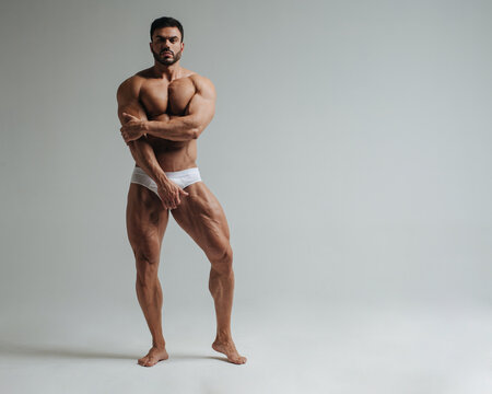 Sexy muscular man in white underwear posing in studio. Handsome bodybuilder standing on grey background. Big male model showing his muscles.