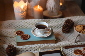 Cup of tea, books, tablet, bowl of cookies, various spices, pine cones and lit candles. Hygge at home. Selective focus.