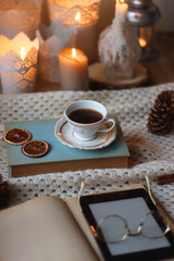 Obraz na płótnie Canvas Cup of tea, books, tablet, bowl of cookies, various spices, pine cones and lit candles. Hygge at home. Selective focus.