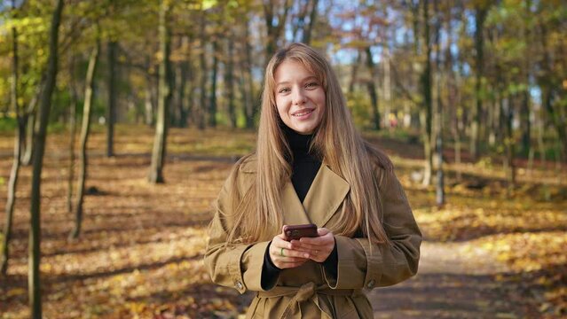 Front view of happy, cheerful lady walking in park in autumn. Pretty, young woman standing, using smartphone, looking at camera, smiling. Concept of harmony with nature.