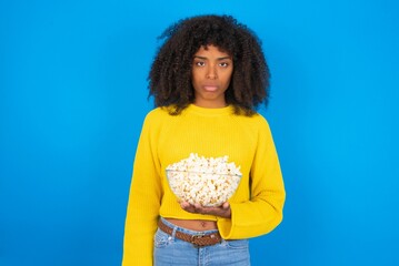 Displeased upset young woman with afro hairstyle wearing yellow sweater over blue frowns face as going to cry, being discontent and unhappy as can't achieve goals,  Disappointed model has troubles