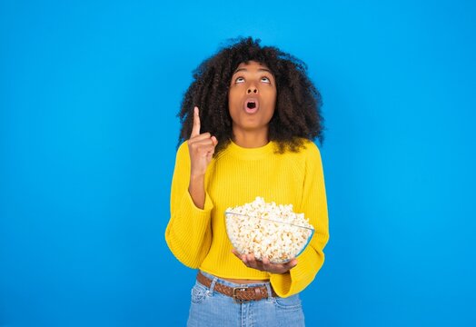 young woman with afro hairstyle wearing yellow sweater over blue wall amazed and surprised looking up and pointing with fingers and raised arms.
