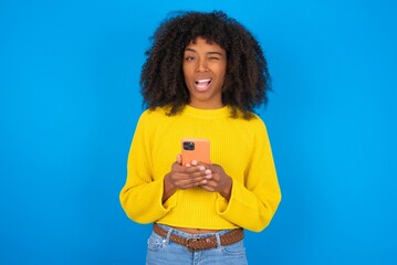 young woman with afro hairstyle wearing orange crop top over blue wall taking a selfie  celebrating success