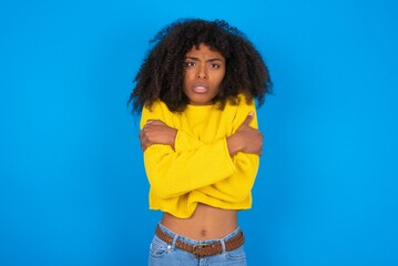 Desperate young woman with afro hairstyle wearing orange crop top over blue wall trembles and feels cold, hugs oneself to warm up or feels scared notices something terrifying.