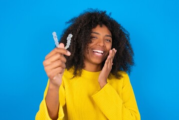 Happy young woman with afro hairstyle wearing orange crop top over blue wall holding and showing at camera an invisible aligner while laughing. Dental healthcare and confidence concept.
