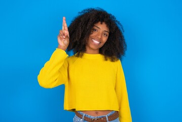 young woman with afro hairstyle wearing orange crop top over blue wall pointing up with fingers number ten in Chinese sign language Shi © Jihan