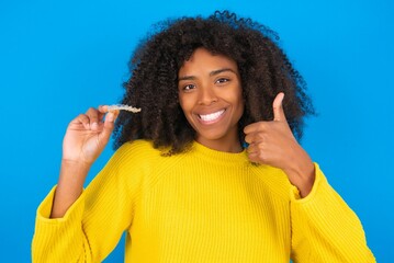 young woman with afro hairstyle wearing orange crop top over blue wall holding an invisible braces aligner and rising thumb up, recommending this new treatment. Dental healthcare concept.