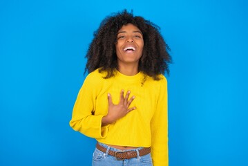 young woman with afro hairstyle wearing orange crop top over blue wall smiles toothily cannot believe eyes expresses good emotions and surprisement