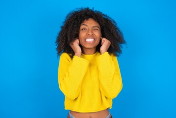 Happy young woman with afro hairstyle wearing orange crop top over blue wall ignores loud music and...