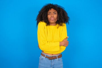 young woman with afro hairstyle wearing yellow sweater over blue wall shaking and freezing for winter cold with sad and shock expression on face.